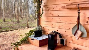 Een houten cabinteinterieur met uitzicht op een bos, een houten plank met diverse voorwerpen, en een kleine tafel met een plant en een zwart object.