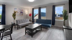 Ein modernes Wohnzimmer mit grauem Sofa, blauem Sofa, Couchtisch und Esstisch mit Stühlen. Es gibt zwei Fenster mit Vorhängen, ein Bild an der Wand und einen Blumentopf.