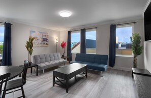Ein modernes Wohnzimmer mit grauem Sofa, blauem Sofa, Couchtisch und Esstisch mit Stühlen. Es gibt zwei Fenster mit Vorhängen, ein Bild an der Wand und einen Blumentopf.