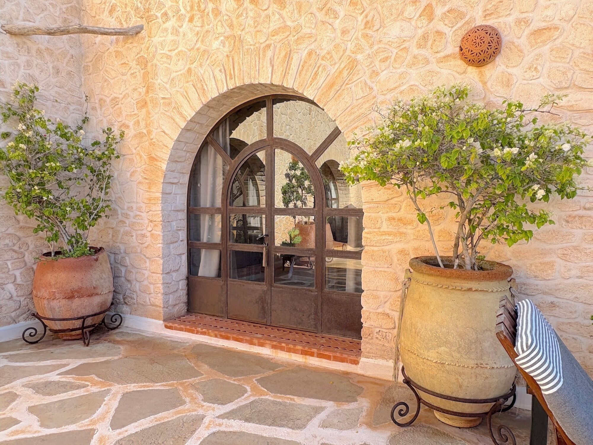 Un portail en pierre en arc de cercle, surmonté d’une grille métallique, avec des plantes en pot et un sol en pierre.