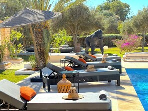 A poolside area with lounge chairs, a statue, and a decorative umbrella. - Palais Tiyana (Ghazoua)