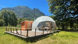 A geodesic dome structure with a wooden deck and a small adjacent building, set in a grassy area with trees and mountains in the background. - Domo Valle El Encanto Peulla (Puerto Varas)