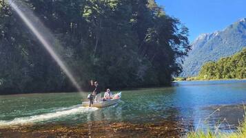 A boat on a calm river with dense forest on one side and mountains in the background.