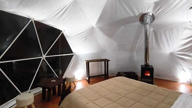 A tented room with a bed, a table, a chair, and a wood-burning stove. - Domo Valle El Encanto Peulla (Puerto Varas)