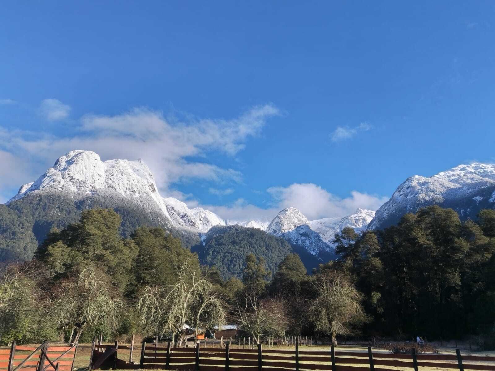 A mountainous landscape with snow-capped peaks, a clear blue sky, and a fenced area with trees.