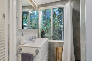 Une salle de bain avec un lavabo blanc, un miroir et une fenêtre donnant sur un paysage verdoyant.