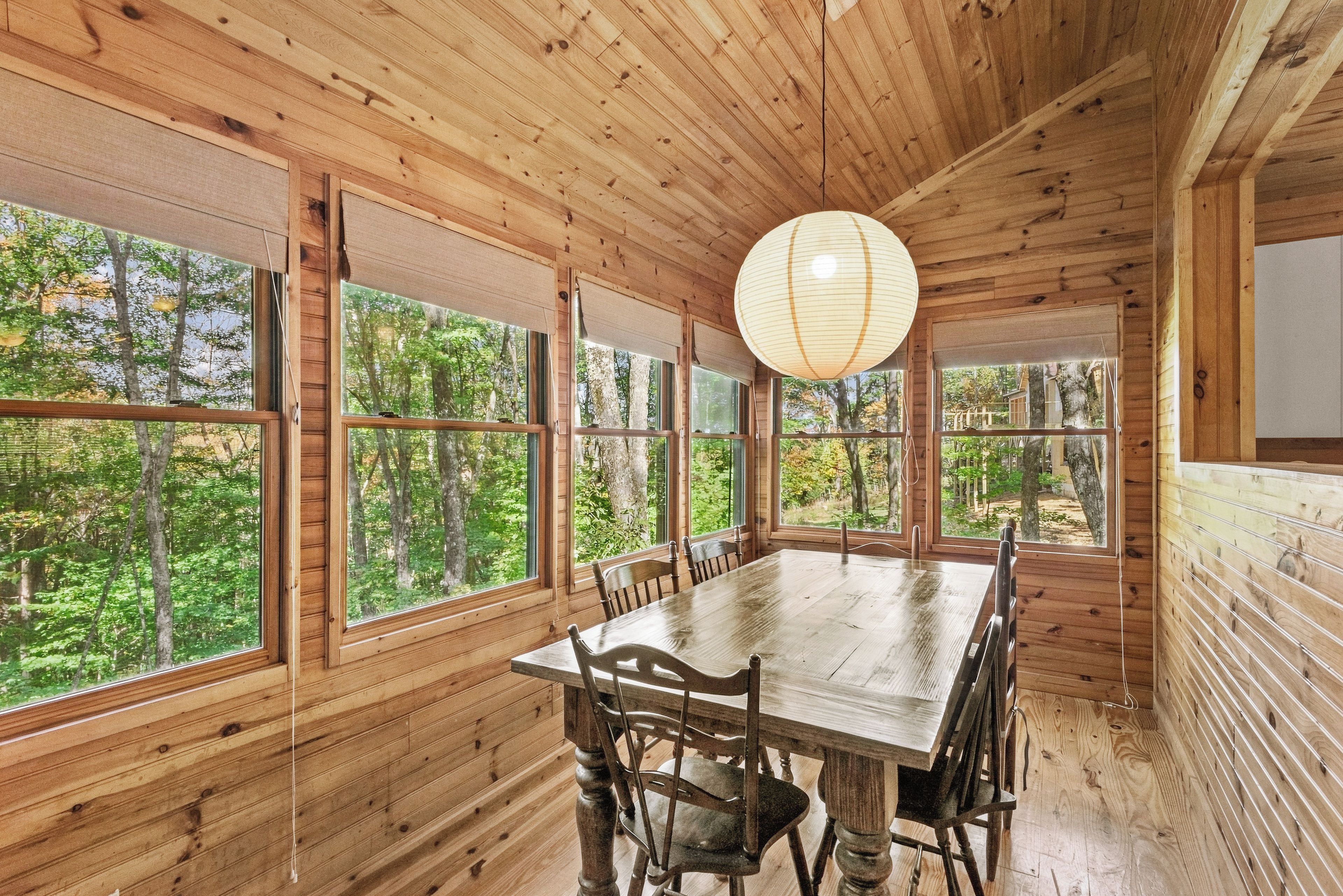 A wooden interior with a dining table and chairs, large windows, and a hanging light fixture.