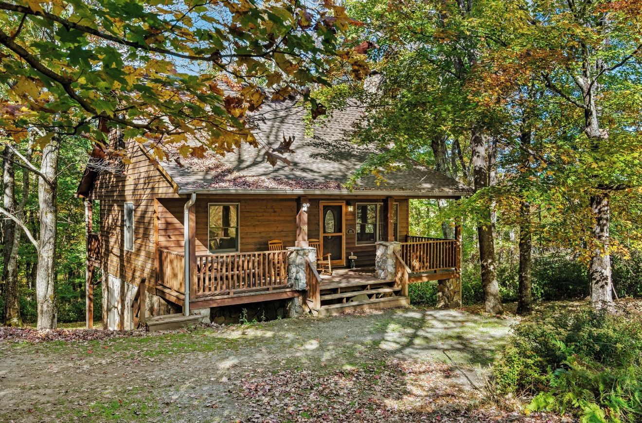 A rustic cabin with a porch, surrounded by trees and a gravel path.