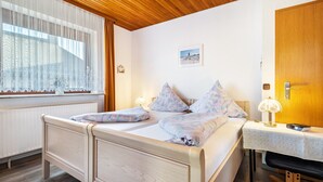 Cama doble con almohadas estampadas, mesita de noche con lámpara, techo de madera y ventana con cortinas de gasa.