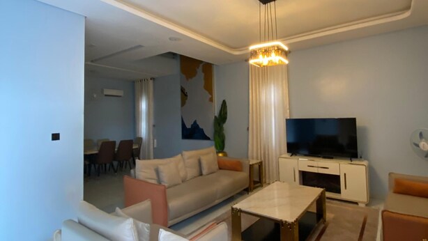 A modern living room with a sofa, coffee table, TV, and a dining area in the background. - ETD SUPPORT (Lekki)