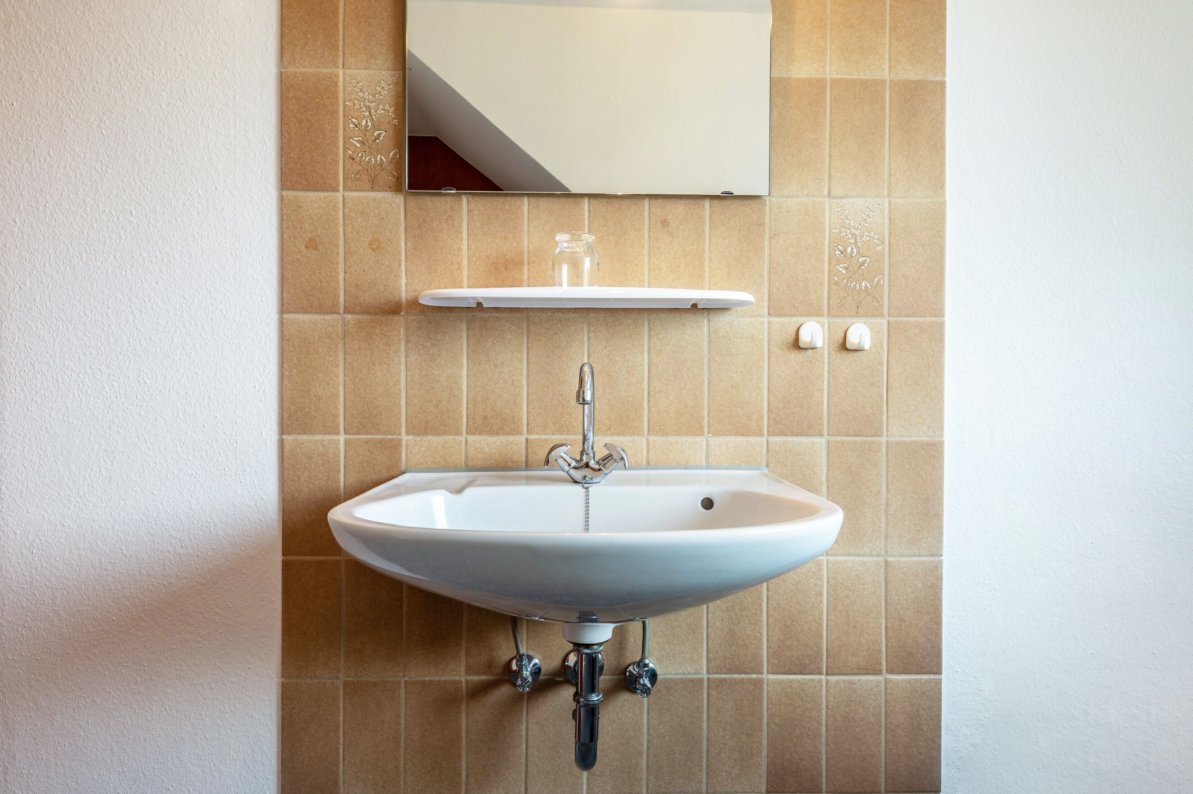 A bathroom with a white sink, a mirror above it, and a shelf with a glass jar.