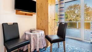 Een kamer met een aan de muur bevestigde televisie, een houten garderobe, een klein rond tafeltje met een schaal en een boek, twee stoelen en een grote schuifglazen deur met uitzicht op bomen.