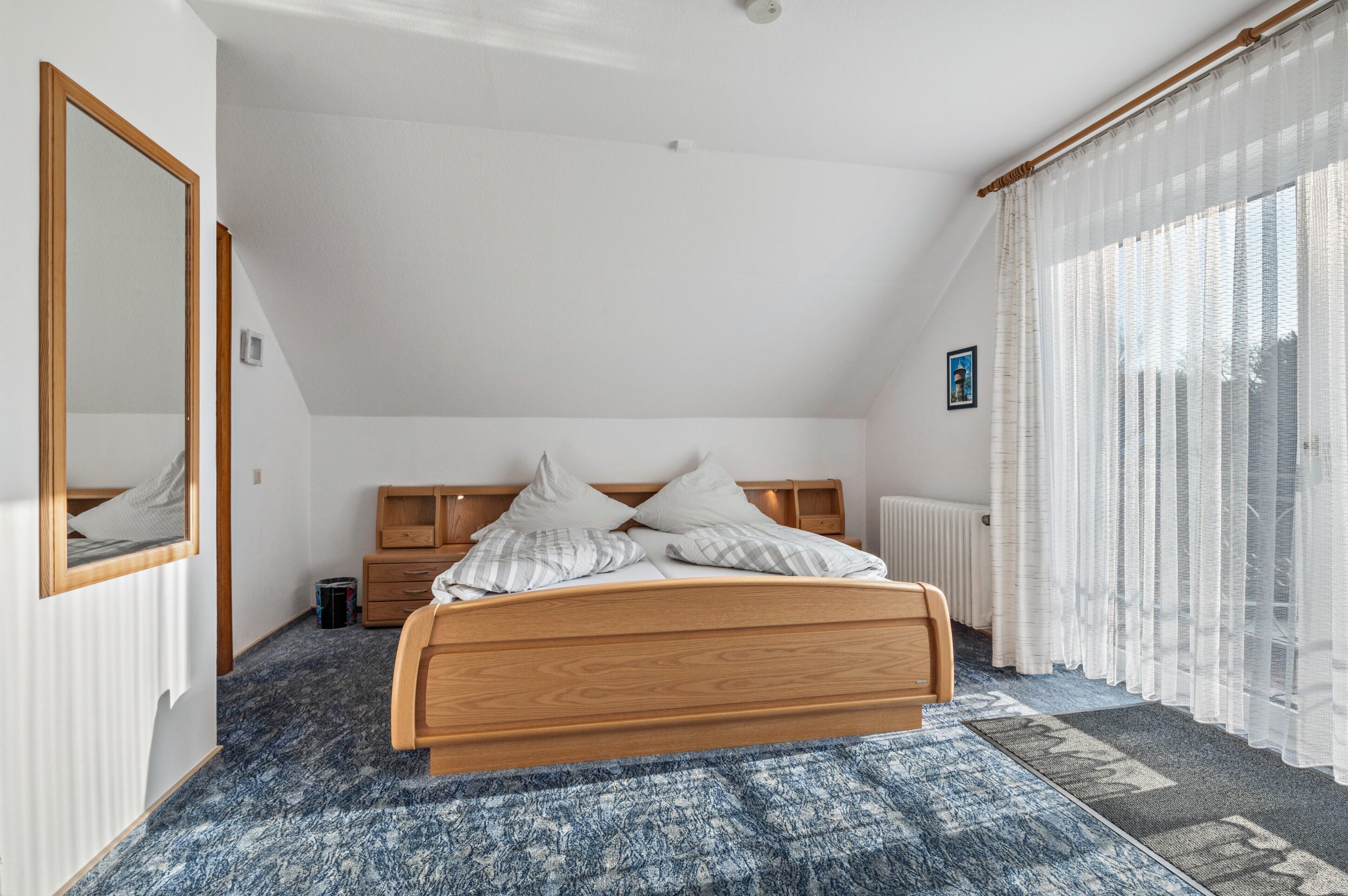 Un dormitorio con cama de madera, un ventanal con cortinas traslúcidas, un radiador y un espejo en la pared.
