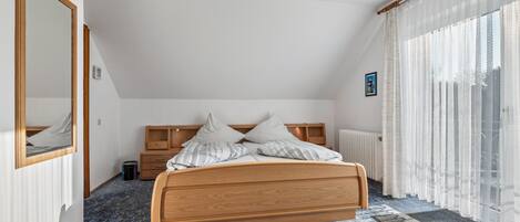 Un dormitorio con cama de madera, un ventanal con cortinas traslúcidas, un radiador y un espejo en la pared.