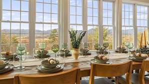 Ein Esstisch, gedeckt für eine Mahlzeit, mit mehreren Weingläsern, Schalen und Tellern, mit Blick auf eine schöne Landschaft durch große Fenster.
