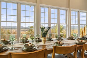 Ein Esstisch, gedeckt für eine Mahlzeit, mit mehreren Weingläsern, Schalen und Tellern, mit Blick auf eine schöne Landschaft durch große Fenster.