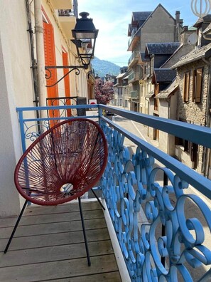 A narrow street with buildings on either side, a blue metal railing, and a red chair on the balcony. - Jasmine, pretty hat. The breath of fresh air. (Bagnères-de-Luchon)