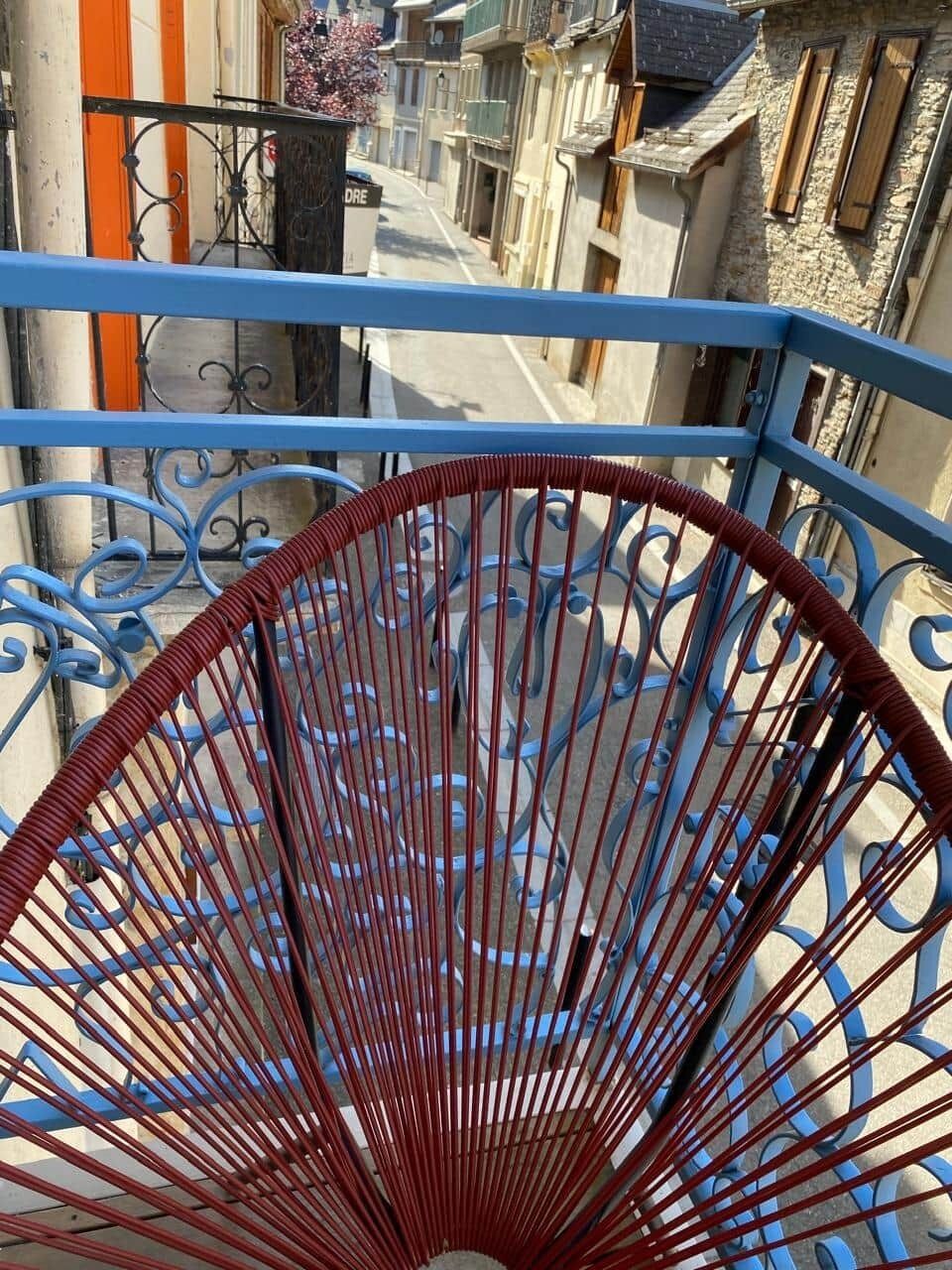 Un escalier en colimaçon avec une rampe bleue et des éléments décoratifs rouges, menant à un espace extérieur où l’on aperçoit des bâtiments en arrière-plan.