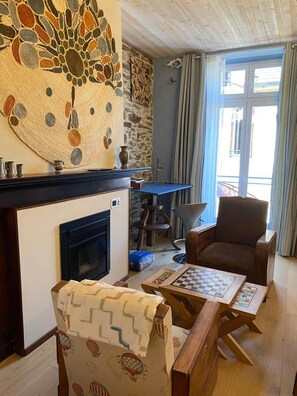 A cozy living room with a fireplace, a chess table, a comfortable armchair, and a large decorative wall piece. - Jasmine, pretty hat. The breath of fresh air. (Bagnères-de-Luchon)