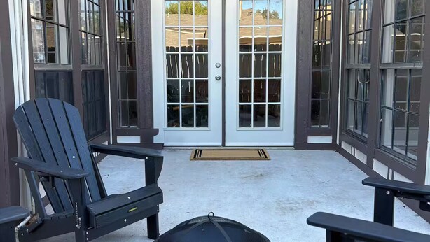En udendørs terrasse med Adirondack-stole og et rundt bord, med udsigt til en bygning med store vinduer og en dobbeltdør.