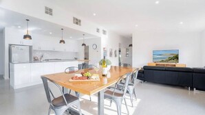 Eine moderne Küche mit weißen Schränken, ein Holzespeisetisch mit Stühlen, ein Fernseher im Wohnbereich und ein großes Fenster mit Meerblick.