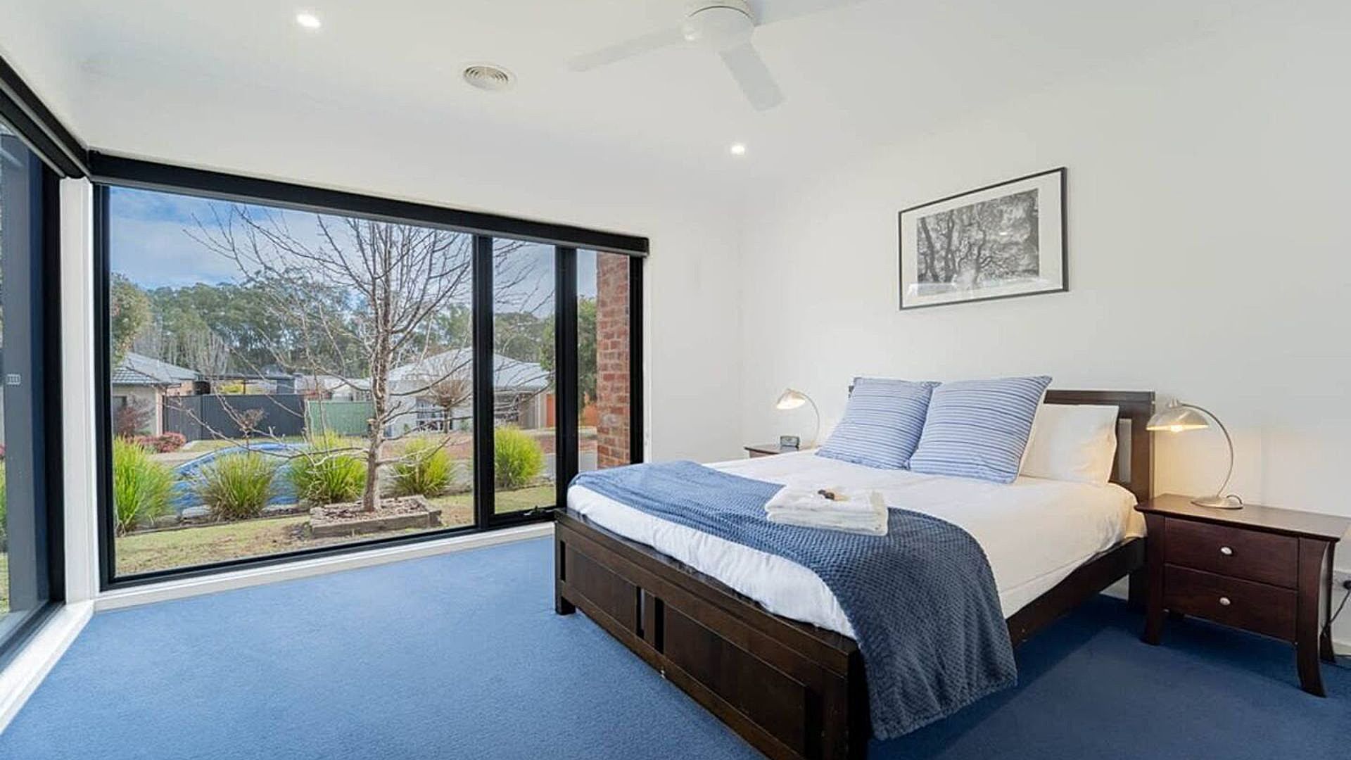 A bedroom with a large sliding glass door leading to a backyard, a bed with a blue and white striped blanket, a bedside table with a lamp, and a framed picture on the wall.