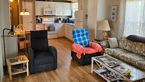 Een ruime woonkamer met een houten vloer, een zwarte fauteuil, een blauwe gestippelde stoel, een bloemenbank en een salontafel met tijdschriften.