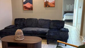 Ein Wohnzimmer mit einem Ecksofa, einem runden Couchtisch und einem grünen Sessel. Zwei Wandbilder zeigen Wüstenlandschaften.