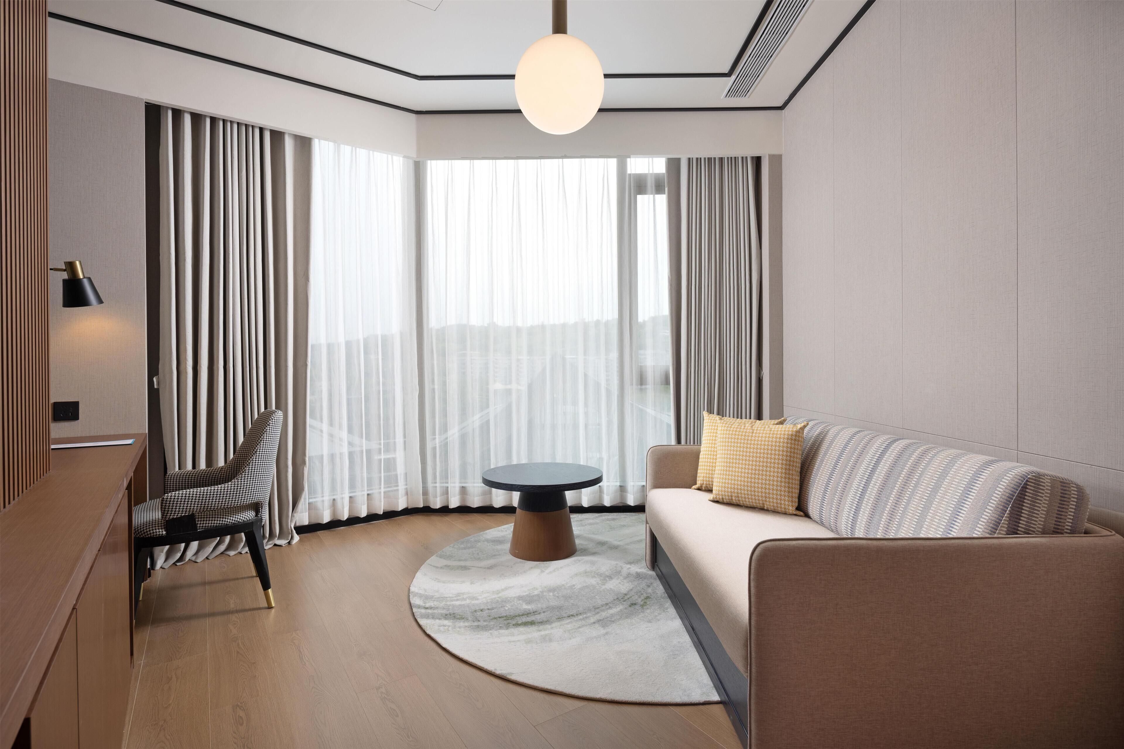 A modern hotel room with a round table, a sofa, a chair, and a large window with curtains.