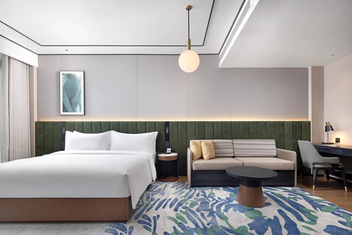A modern hotel room with a large bed, a sofa, a desk, and a patterned rug.