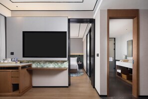 A modern hotel room with a flat-screen TV, wooden furniture, a bathroom with a mirror, and a hallway leading to another room. - Hilton Garden Inn Chongqing Bishan (Chongqing)