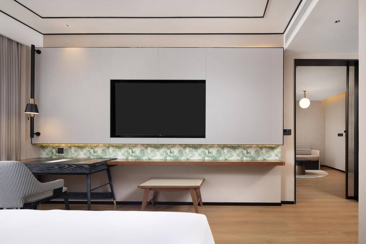 A modern hotel room with a flat-screen TV mounted on the wall, a small wooden table, a chair, and a bed.
