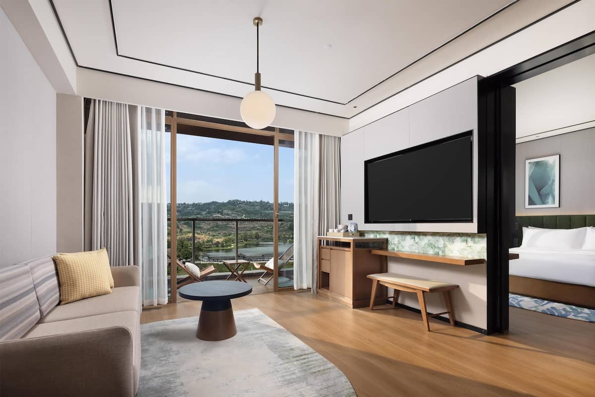 A modern hotel room with a large TV, a sofa, a dining table, and a balcony with a view.