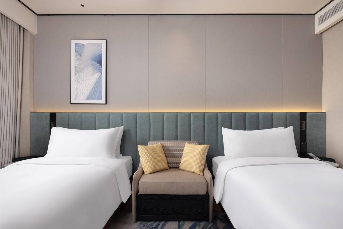 A hotel room with two beds, a small seating area, a framed artwork on the wall, and a ceiling-mounted air conditioning unit.