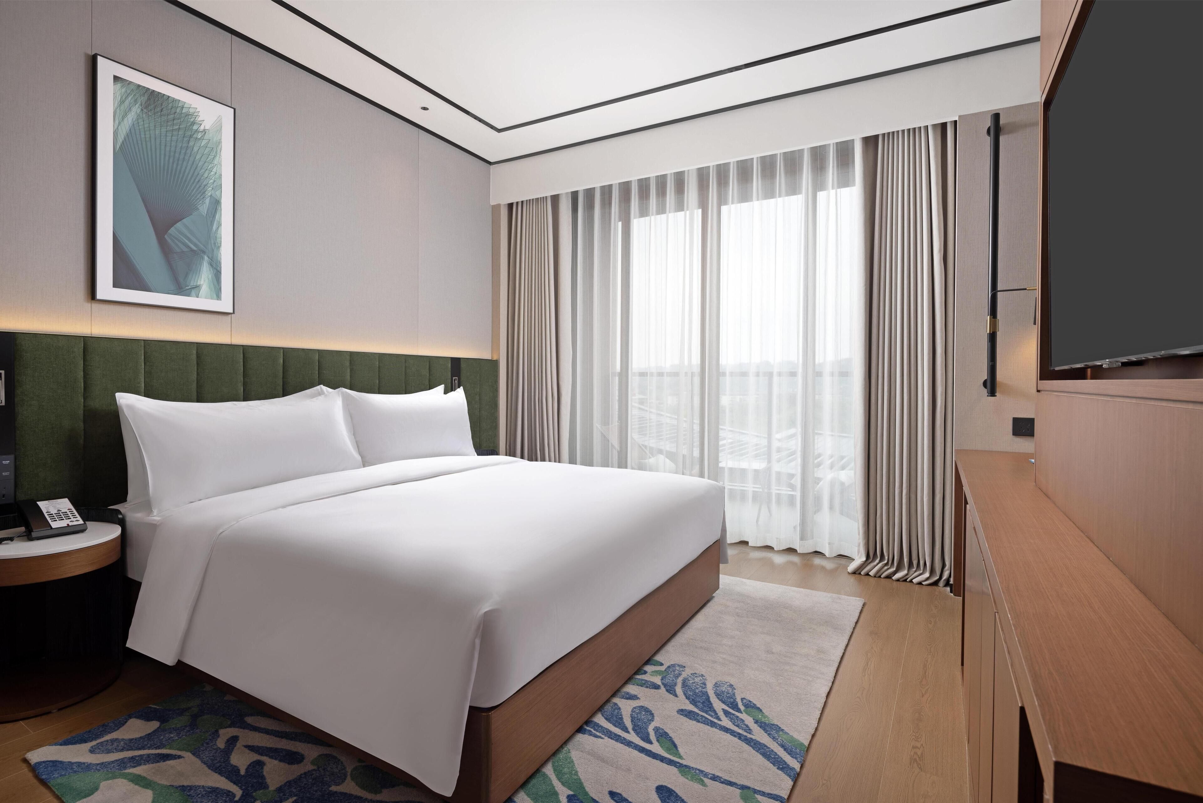 Une chambre d’hôtel avec un lit, une table de chevet, une télévision et une grande fenêtre avec des rideaux.