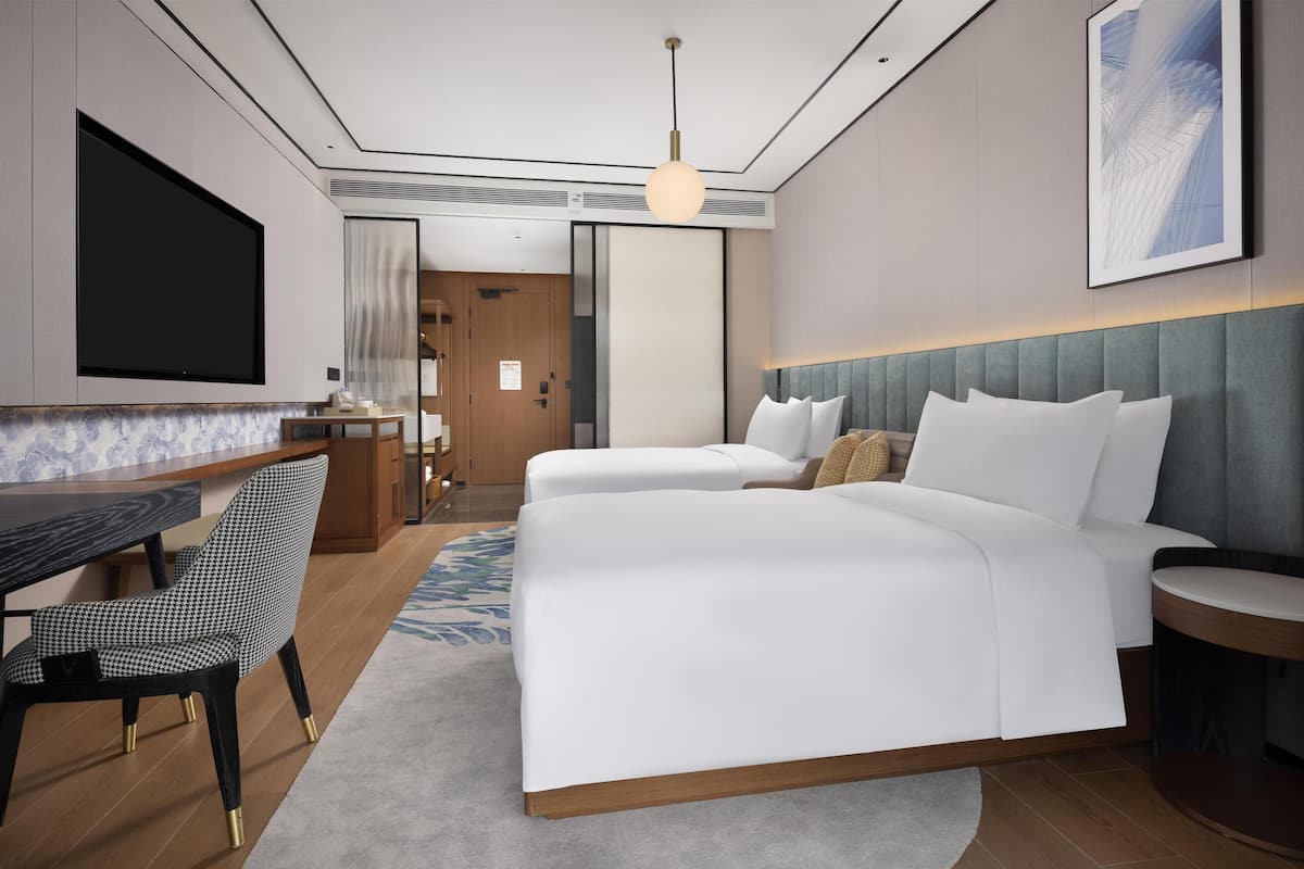 A modern hotel room with a large bed, a desk with a chair, a flat-screen TV, and a large window.
