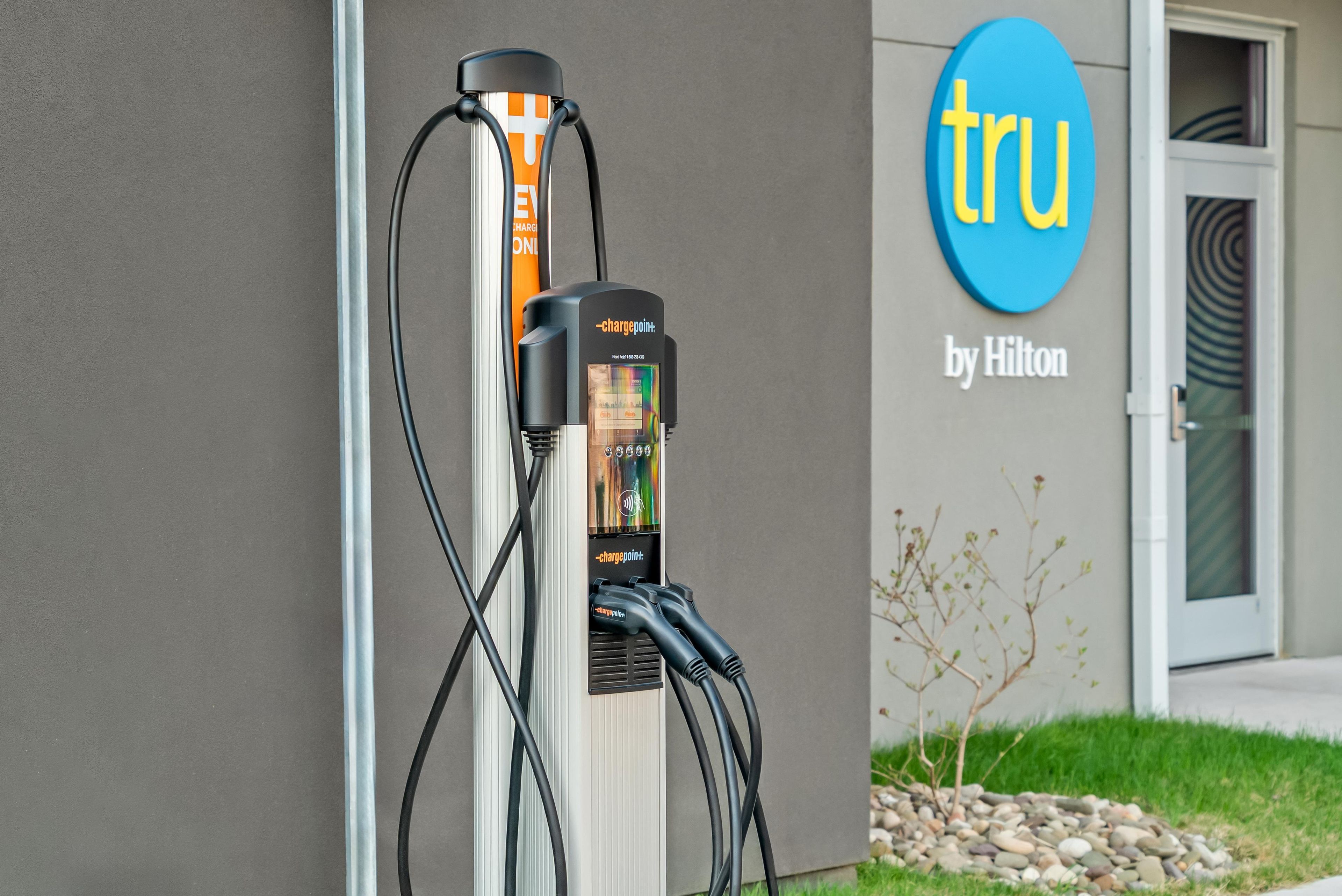 An electric vehicle charging station with a digital display, connected to a building with a 'tru by Hilton' sign.