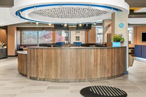 A hotel reception area with a curved wooden reception desk, a large window with a view of trees, and a blue sign indicating the hotel name. - Tru By Hilton Hagerstown (Hagerstown)