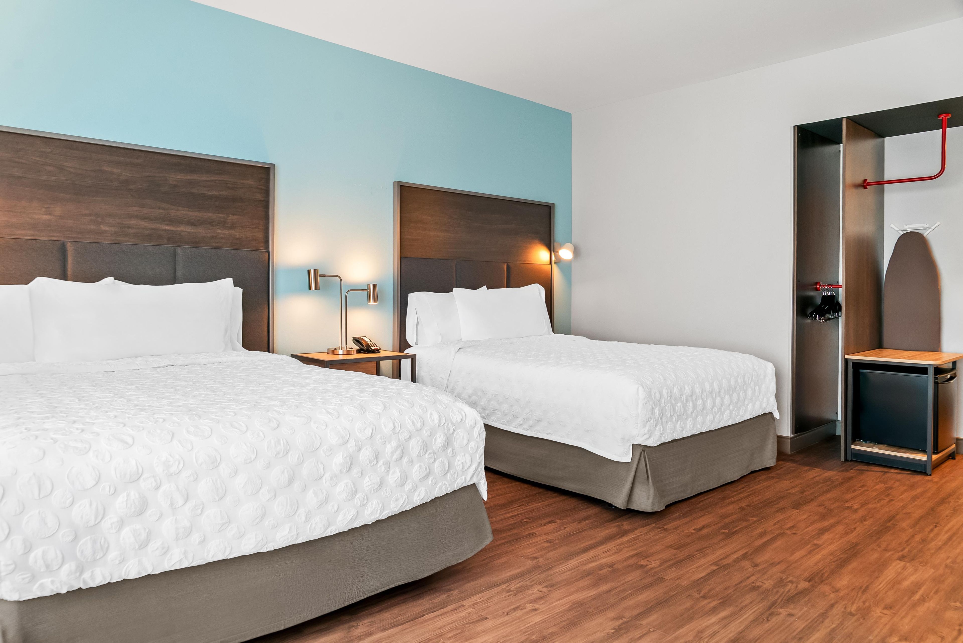 A hotel room with two beds, a nightstand, a wall-mounted lamp, and a door with a towel rack.