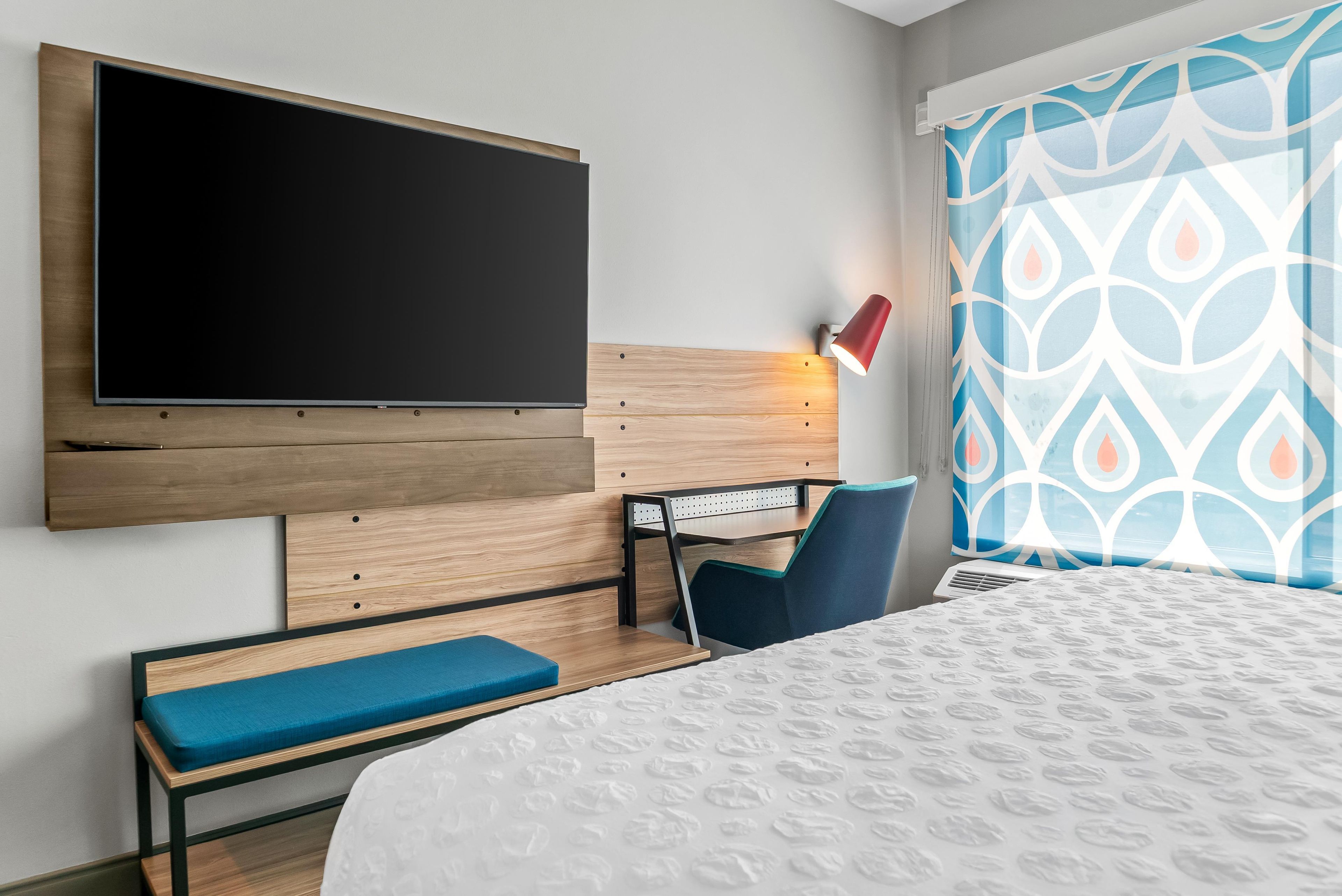 Camera d'albergo con TV a schermo piatto montata a parete, comodino con lampada, una sedia blu, una panca e un letto con copripiumino a fantasia.