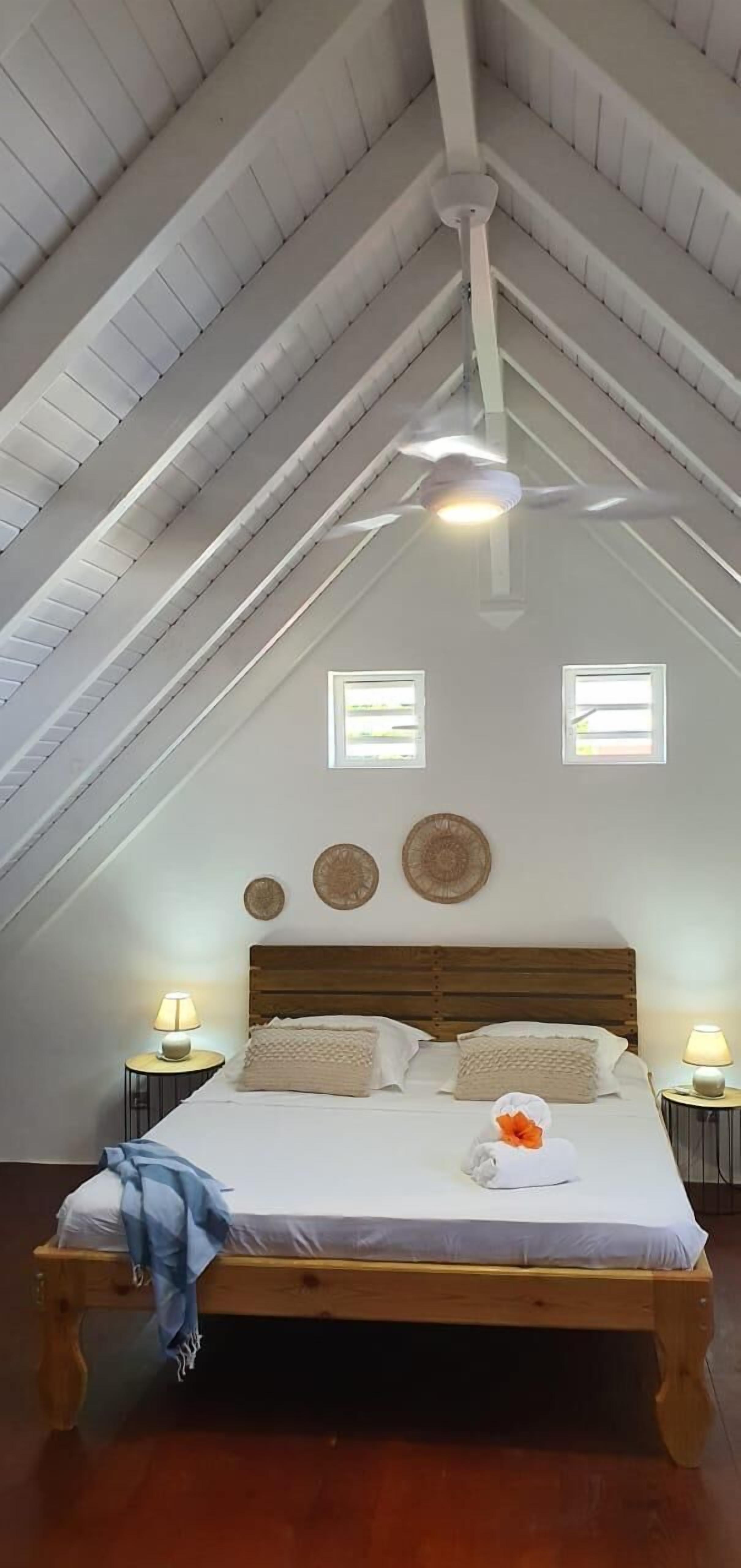 Une chambre avec un plafond en pente, un lit en bois, deux tables de chevet avec des lampes et deux petites fenêtres.