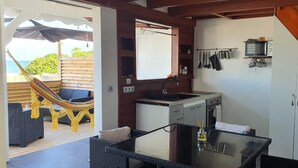 Une cuisine compacte équipée d’un petit évier, d’un four à micro-ondes et d’une cuisinière. On aperçoit un hamac et un espace extérieur aménagé pour s’asseoir à travers une porte ouverte.