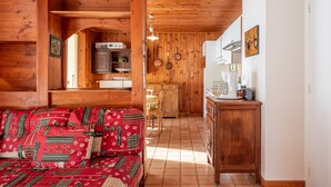 A cozy cabin interior with a wooden ceiling, a sofa with patterned cushions, a kitchen area with wooden cabinets, and a dining table.