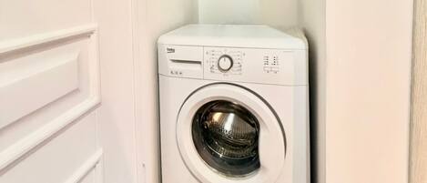 Una lavadora blanca y compacta en un lavadero pequeño con piso de madera y paredes blancas.