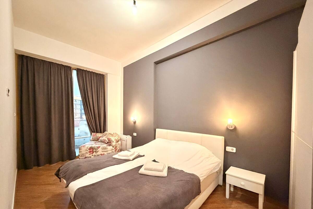 Un dormitorio con una cama grande, dos mesitas de noche, una silla y una ventana con cortinas.