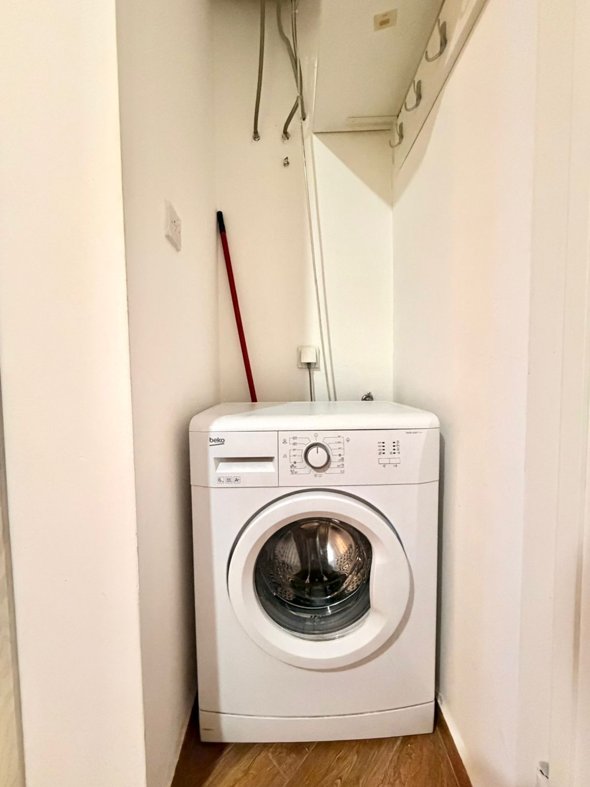 A compact, white front-loading washing machine in a narrow hallway with wooden flooring.