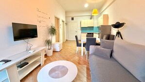 Un salon moderne avec un téléviseur à écran plat, une table basse blanche, un canapé gris et un coin repas avec une cuisine en arrière-plan.