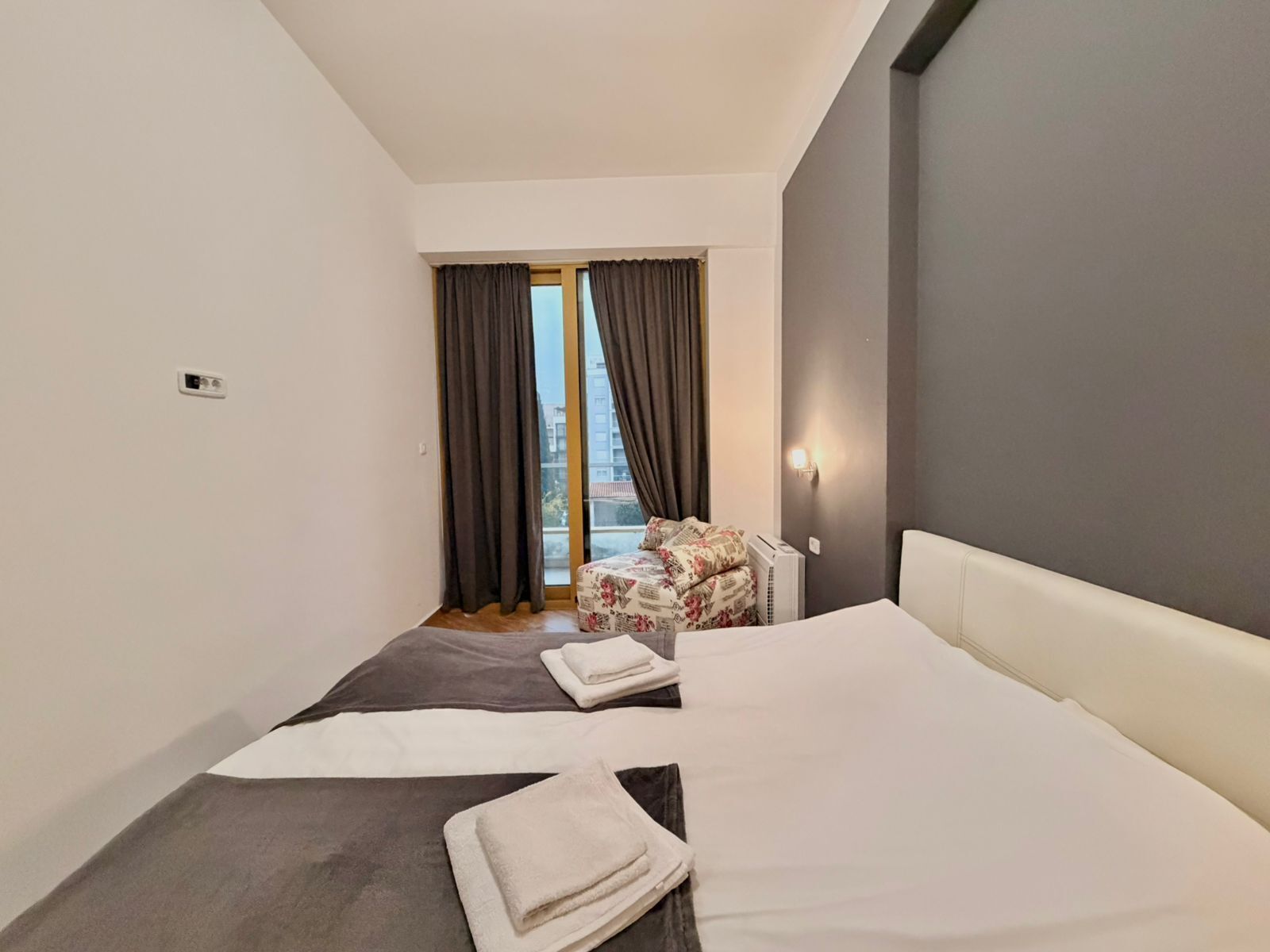 A hotel room with a large bed, a small seating area with a chair and a table, a window with curtains, and a wall-mounted light fixture.