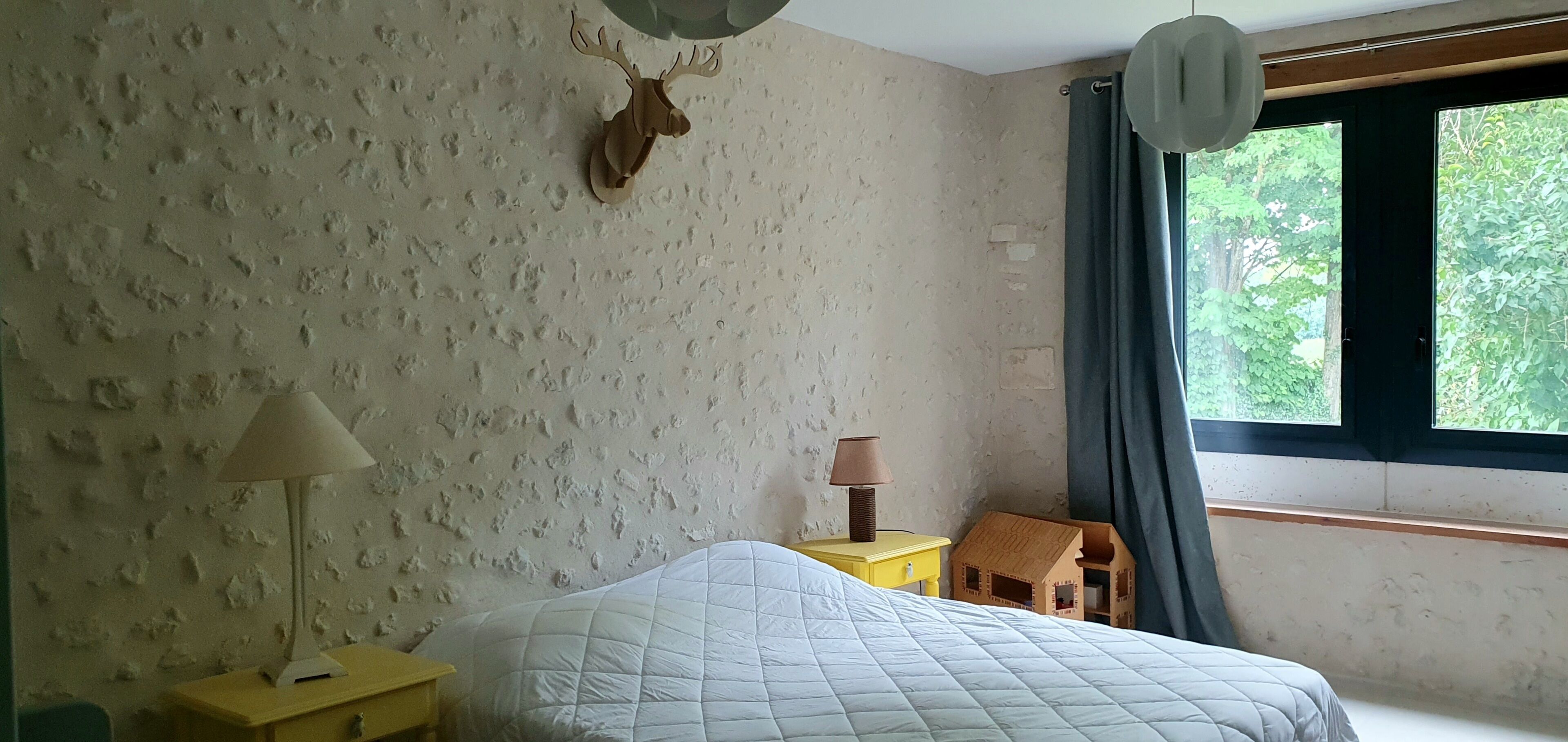 A bedroom with a bed, a lamp, a small wooden table, a decorative deer head, and a window with a view of greenery.