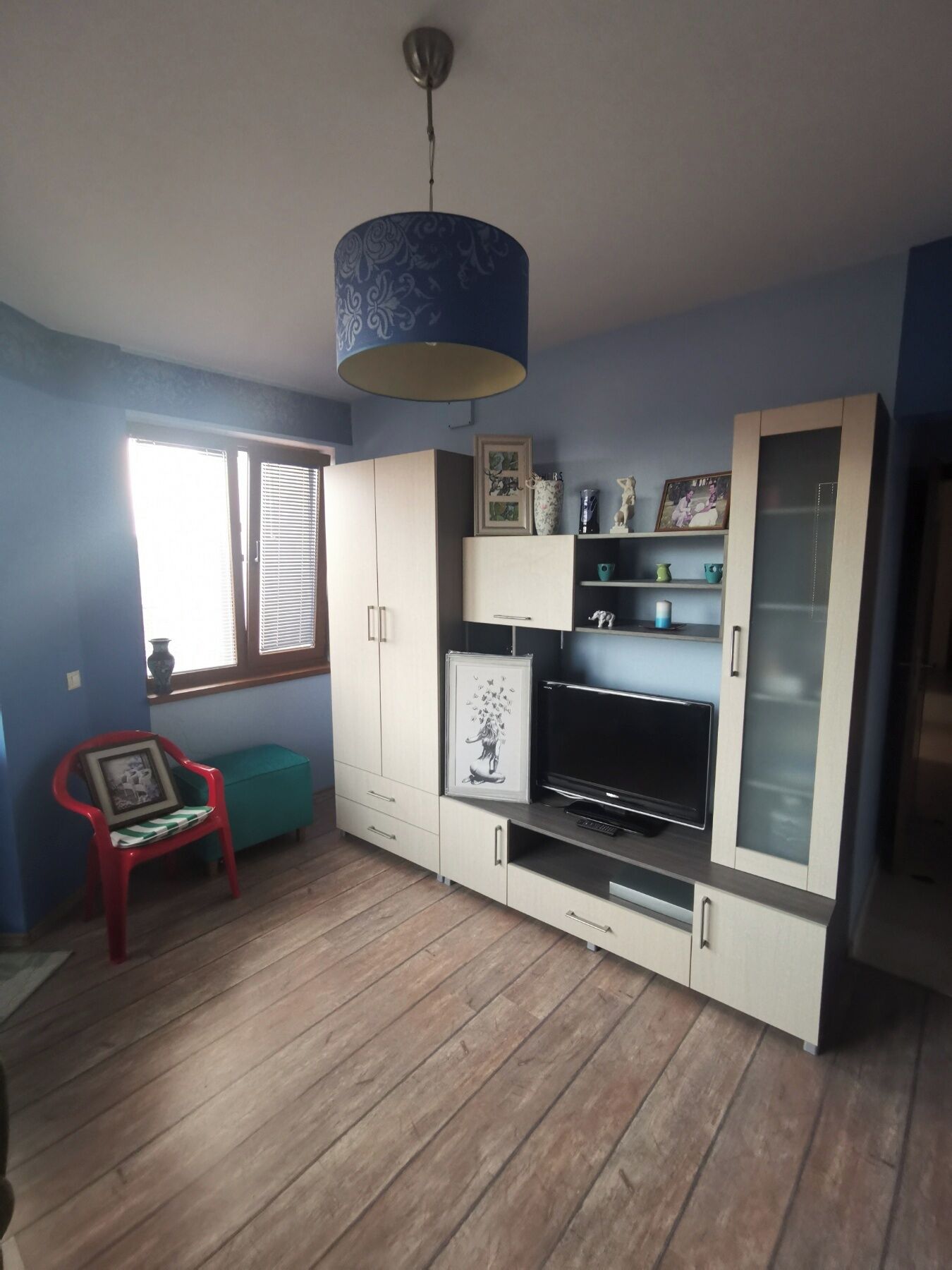 Ein Wohnzimmer mit Holzboden, blauer Wand, einem TV-Schrank mit Flachbildfernseher, einem roten Sessel und einem Deckenventilator.